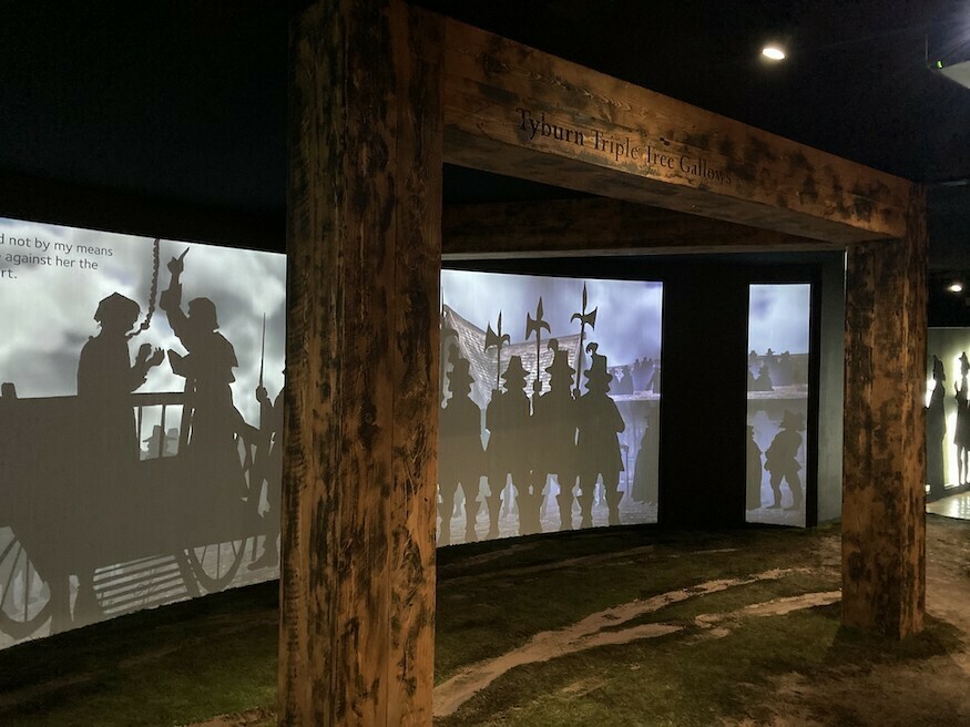 A large wooden gallows with three legs, with projected images of executions in the background