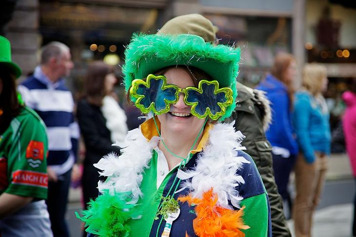 A woman with shamrock shaped comedy glasses, a green furry hat, and a green, white and orange feather boa, at the St Patrick's Day parade