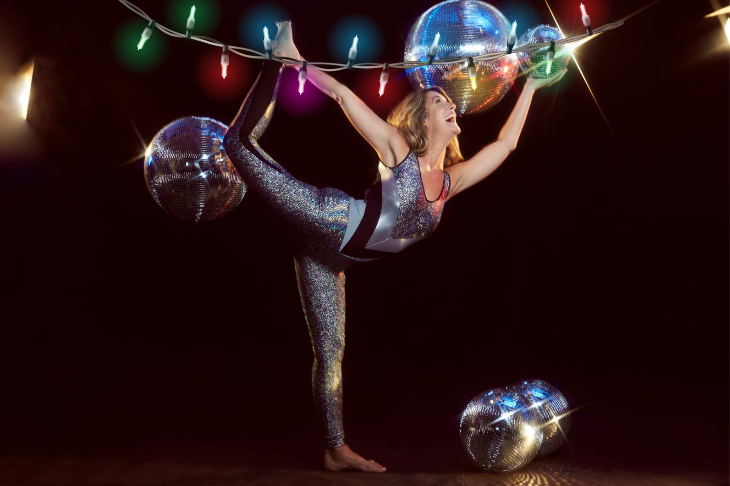 A woman in sparkly workout clothes pulling a yoga pose, surrounded by disco balls and Christmas lights