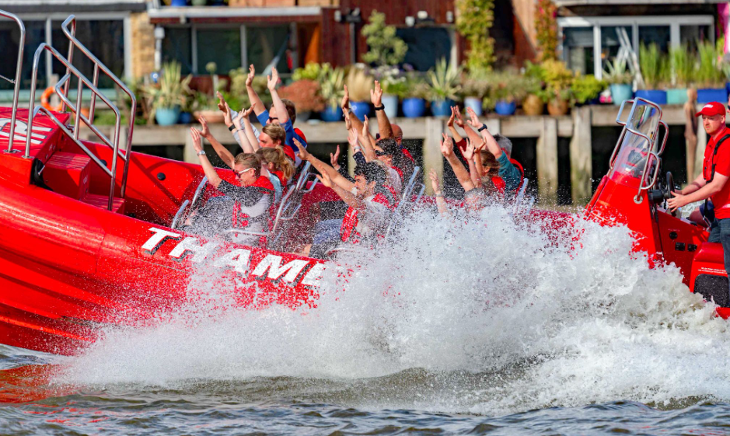 People sitting in a moving Thames Rockets red speedboat, holding their hands in the air while spray splashes them