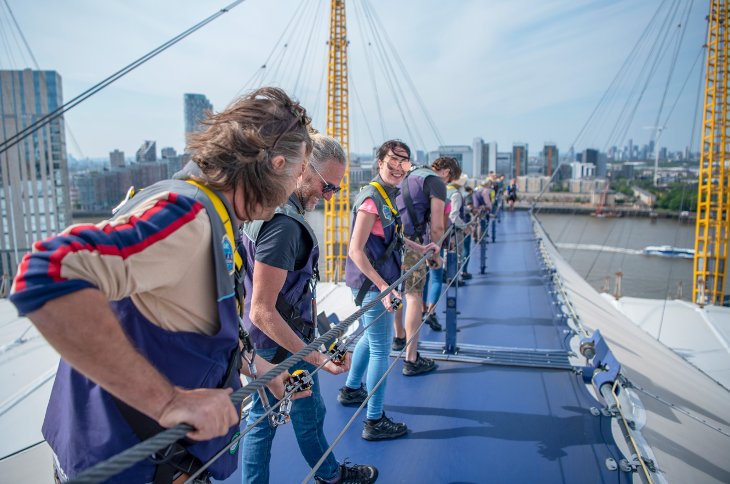 A group of people in a row holding onto a railing and walking on a blue path over the O2