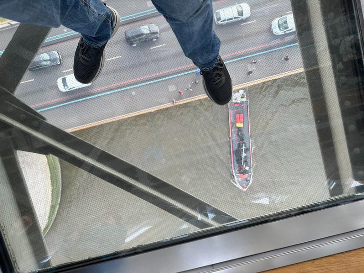 A pair of feet in black trainers standing on a glass floor, with the Tower Bridge road, and a  boat on the river visible below