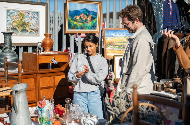 A couple looking at a small glass vase, surrounded by other flea market items