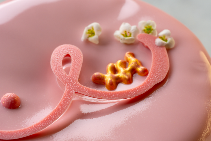 A pink iced pastry decorated with an abstract representation of two breasts, one with a golden squiggle